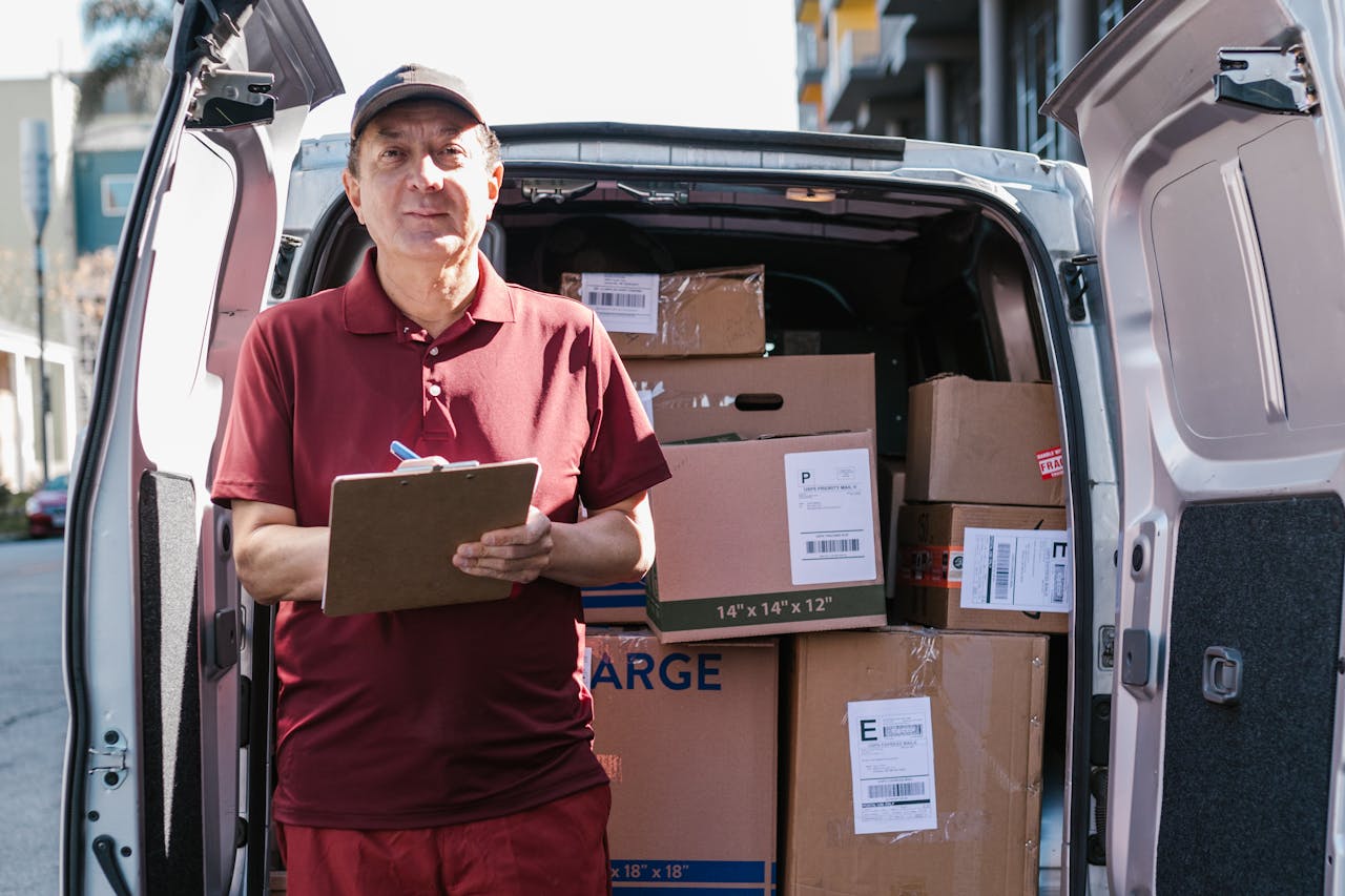 about-us Elderly delivery person holds clipboard in front of open van filled with packages on a sunny day.